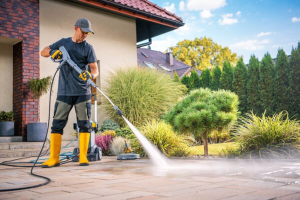 A man is using a power washer to clean the patio in a beautiful garden filled with greenery and sunshine.